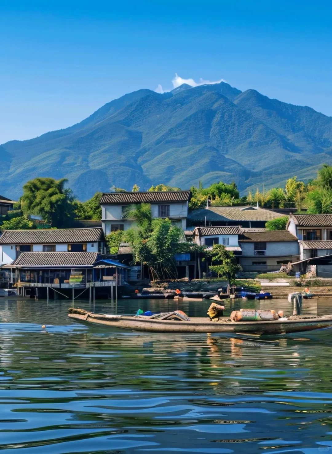 Yunnan travel recommendation — Erhai Lake fishing village in Dali, a wooden cargo boat moored on the calm lake surface, white-walled grey-roofed Bai-style houses and bamboo groves behind, Cangshan Mountain peaks rising in the distance, clear blue sky reflected on the water