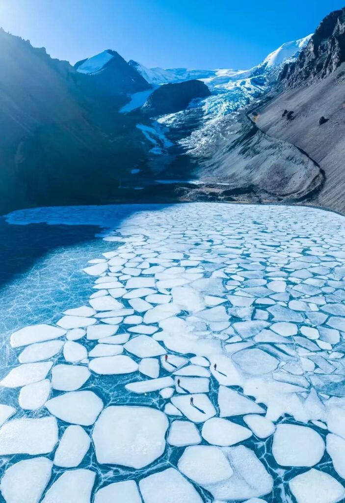 恰央措冬季冰湖航拍,湖面結成大片白色冰塊拼接圖案,背景為冰川雪山,遊人站於冰面顯出壯闊尺度