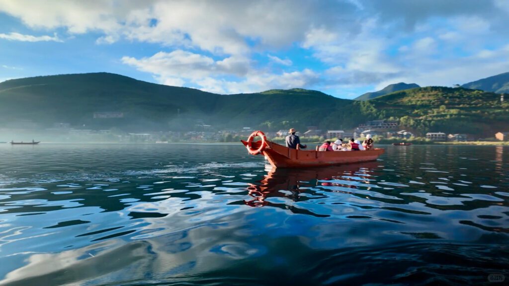 雲南旅遊推薦|昆大麗瀘沽湖8日深度慢遊,最值得的雲南行程 - CITS China International Travel Service 一艘载着数名游客的朱红色木船行驶在清晨的泸沽湖湖面上,船夫戴斗笠划桨,湖面倒映蓝天白云,远岸薄雾缭绕,翠绿山坡与湖滨村落依稀可见