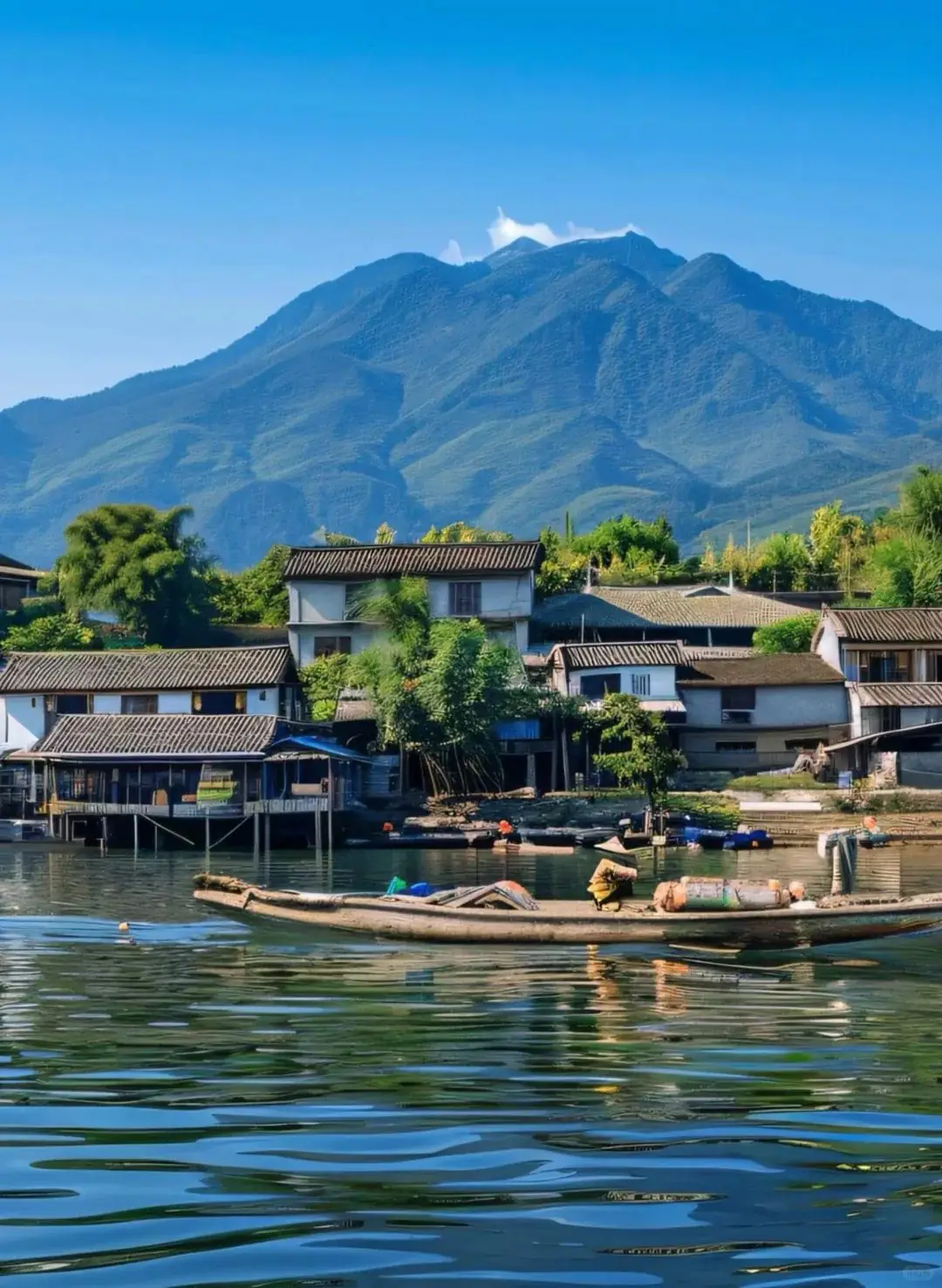 雲南旅遊推薦.大理洱海岸邊漁村聚落景觀，一艘載貨木船停泊於平靜湖面，背後是白牆灰瓦民居與翠綠竹林，遠處蒼山青峰高聳，藍天無雲，湖面倒映山色