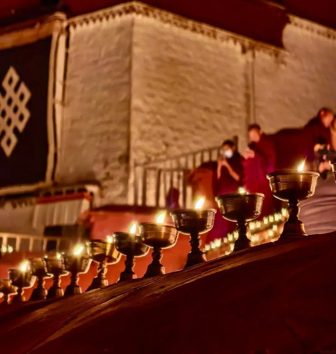 Nighttime lamp offering at Ganden Monastery during the Tibetan Festival of Lights; a row of butter lamps in the foreground, with a monk lighting a lamp beside the monastery wall